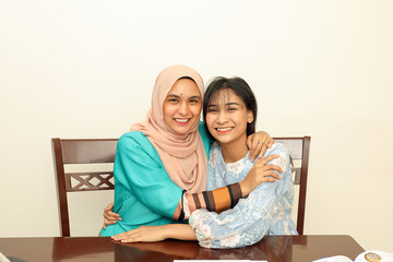 Two young Asian Malay Muslim woman wearing headscarf at home office student sitting at table phone computer book document selfie self portrait hug care affection