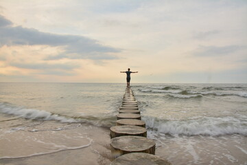 A young boy standing on stilts in the sea with his arms outstretched, looking at the sunet © TOMASZ