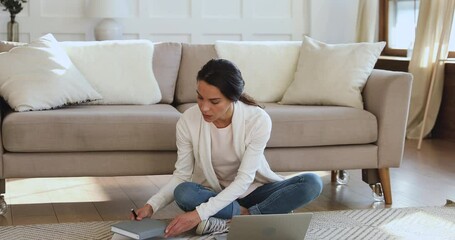 Focused young biracial female student sitting on floor with computer, doing homework alone indoors. Concentrated millennial mixed race freelance woman organizing workday in paper planner at home.