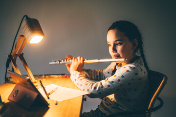 Remote music lesson. Girl playing the flute at home at the night.
