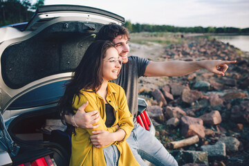 Happy traveler couple sitting in car open trunk and watch the sunrise