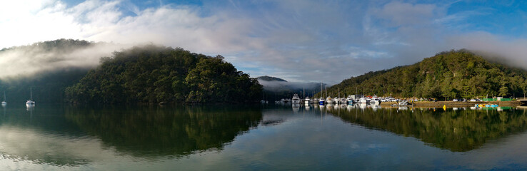 Fototapeta na wymiar Beautiful morning panoramic view of Cockle creek with reflections of blue sky, light clouds, foggy mountains and trees, Bobbin Head, Ku-ring-gai Chase National Park, New South Wales, Australia