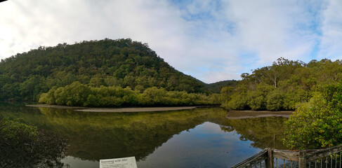 Fototapeta premium Beautiful morning view of Cockle creek with reflections of blue sky, foggy mountains and trees, Bobbin Head, Ku-ring-gai Chase National Park, New South Wales, Australia