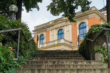 Treppe mit Blick auf die Fassade des Festspiel Gebäudes - Wagner Festspielhaus Bayreuth. Orangene Steine, Blaue Fensterrähmen und Sandsteine. Bäume und Treppe. Bayreuth in Bayern, Deutschland. 
