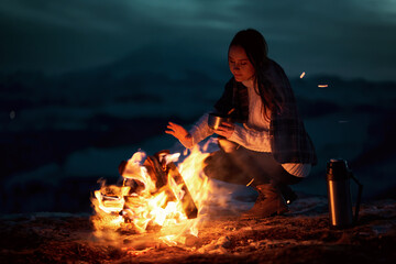 The girl traveler in the evening warms himself by the fire in the mountains near the camp, the girl is lit by the light of fire. Against the backdrop of a blurred mountain range and Elbrus volcano