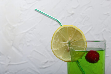 Green lemonade in a glass. Cherry floats in it. Garnished with a slice of lemon. With a straw for a cocktail.