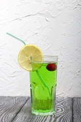 Green lemonade in a glass. Cherry floats in it. Garnished with a slice of lemon. With a straw for a cocktail.