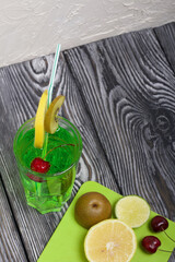 Green lemonade in a glass. Cherry floats in it. Garnished with a slice of lemon and kiwi. With a straw for a cocktail. Nearby on a cutting board are fruits for a cocktail.
