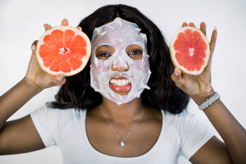 Joyful smiling young African girl with facial sheet mask, posing to camera on white background with...