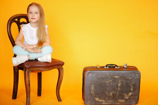 Little young blonde Girl sitting on old vintage chair in yellow studio and smiling next to old vintage suitcase travelling trunk . Travaller style concept. Voyager adventure lifestyle, Travel style