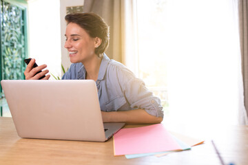 a woman working on her computer who is on the cell phone and who is teleworking at home