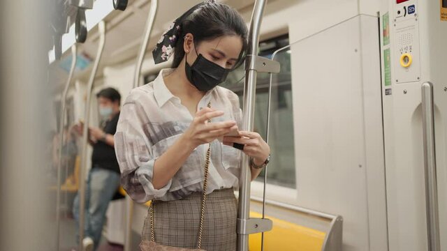 Beautiful Asian Lady Wear Mask Stand Hold On To Pole Inside Subway Car Head Down Using Cellphone, Commuter On The Ride, Covid-19 New Normal, Risk On Public Transport, Social Distance, Smartphone User