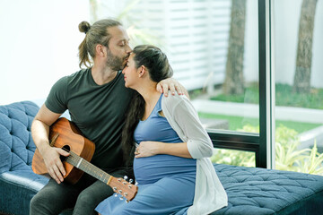 Man hold guitar and kiss on his pregnant wife forehead to express love and care in family. Concept of take care and support lover people in parent also stay at home to protect from virus pandemic.