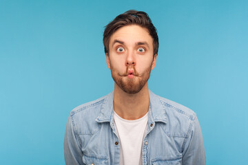 Obraz premium Portrait of humorous funny man in worker denim shirt making amazed grimace with comical fish face, pout lips and surprised silly idiotic expression. indoor studio shot isolated on blue background