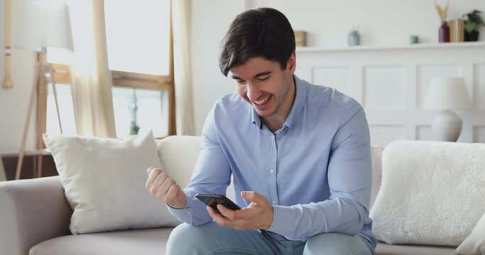 Smiling young handsome guy sitting on sofa, looking at cellphone screen, celebrating online lottery win notification. Overjoyed happy millennial man received message with good news on smartphone.