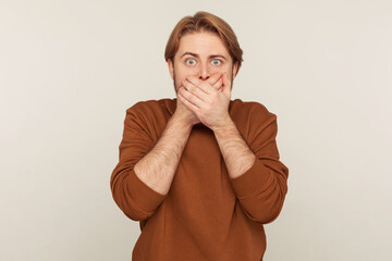 I won't speak! Portrait of terrified young man in sweatshirt covering mouth and looking scared...