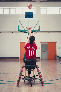 Female Chinese Paralympic Athelte In Wheelchair Shooting Basketball Ball Practice In Gym.