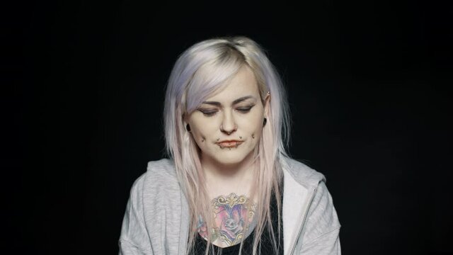 Close up of woman with white hair and tattoo on body with her eyes closed. Woman with fashion piercing on lips and cheeks on black background.
