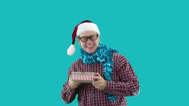 A man dressed in a plaid shirt and glasses on his head holds a New Year's cap in his hands a New Year's box with a gift and a Christmas ball, smiles and teases to give or not to give.