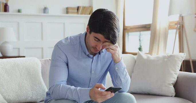 Frustrated young man looking at smartphone screen, feeling disappointed with received bad news message. Stressed millennial guy reading sms, frustrated by annoyed bank loan notification indoors.