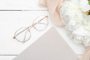Flat lay composition with elegant female eyeglasses, paper notebook, beige fabric and peonies on white wooden table. Rustic style, view from above.