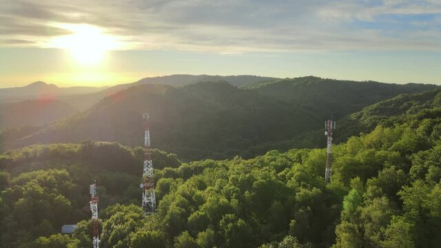Aerial over green mountains with 5G telecommunication towers standing tall against beautiful sunlight.