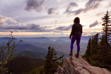 Fototapeta premium Adventurous Girl na szczycie Rocky Mountain z widokiem na piękny kanadyjski krajobraz przyrody podczas dramatycznego zachodu słońca. Zrobiono w Chilliwack, na wschód od Vancouver w Kolumbii Brytyjskiej w Kanadzie.