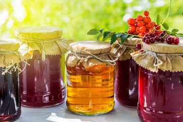 Fruit and berry preserves in the glass jars and raw strawberries , cherries , rowans and red currants berries on a white wooden table, on the nature background, organic meal and dessert concept