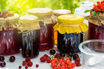 Fruit and berry preserves in the glass jars and raw strawberries , cherries , rowans and red currants berries on a white wooden table, on the nature background, organic meal and dessert concept