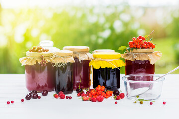 Fruit and berry preserves in the glass jars and raw strawberries , cherries , rowans and red currants berries on a white wooden table, on the nature background, organic meal and dessert concept