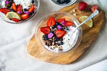 Yogurt with granola, bilberries, strawberries, seeds, pine nuts and sweet William flowers in glass bowl on wooden board on white background.  