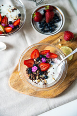Yogurt with granola, bilberries, strawberries, seeds, pine nuts and sweet William flowers in glass bowl on wooden board on white background.  