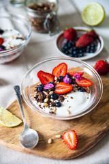 Yogurt with granola, bilberries, strawberries, seeds, pine nuts and sweet William flowers in glass bowl on wooden board on white background.  