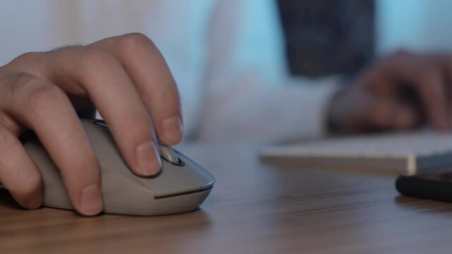 Male hand clicking on grey mouse and typing on a keayboard, gimbal shot. Male hand on a white mouse on blurred background of man dressed in white shirt, no face
