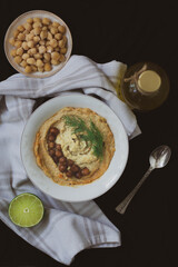 Chickpea vegan hummus garnished with dill and baked chickpeas on black background with vintage spoon, bottle of olive oil, towel, chickpeas and half a lime