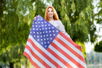 Happy young woman posing with USA national flag standing outdoors in summer park. Positive girl with United States banner outdoors.