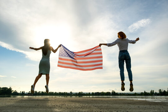 Two young friends women holding USA national flag jumping up together outdoors at sunset. Patriotic girls celebrating United States independence day.
