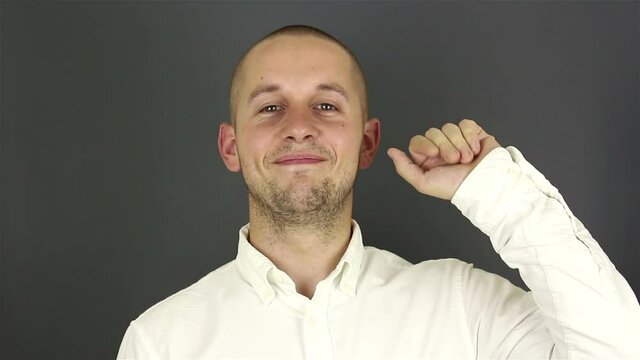 Young handsome guy in a white shirt, says goodbye and waves his hand. Portrait on a gray background.