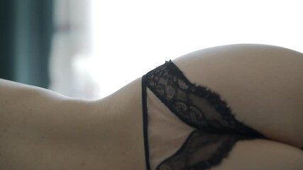 Back of attractive young woman in black sexy lingerie lying on bed close up, beautiful female body in lace panties against window with curtain moved by the wind.
