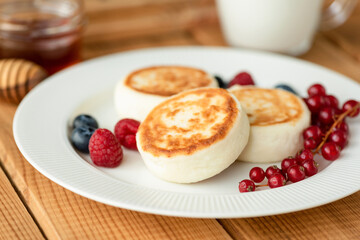 Syrniki cottage cheese fritters with summer berries on white plate, wooden table background. Russian sweet breakfast cheese pancakes