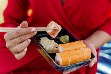 Close-up sushi set. Attractive girl in red dress holds sushi with chopsticks in hands. Courier delivered sushi set for pretty woman. Black box with sushi, wasabi, ginger.