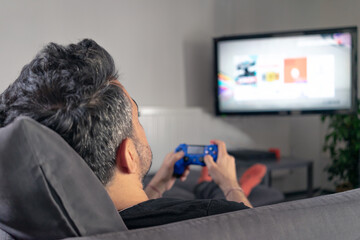 man playing video game at home sitting on coach sofa in front of television with play game concole and joystick in hands