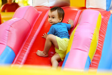 Toddler boy in blue shirt and yellow shorts climbing up slide on bright inflatable bouncy castle. Little kid half turned on bouncing attraction at playground, maybe he is looking for parents attention