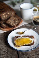 Slice of homemade traditional Danish sourdough rye bread with butter and honey on white and blue plate, cup of milk, coffee and bread loaf on wooden background.