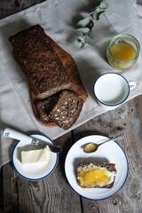 Homemade traditional Danish sourdough rye bread. Sliced bread on wooden board, cup of milk, butter and bread slice  with butter and honey on white and blue plates and honey jar on wooden background.