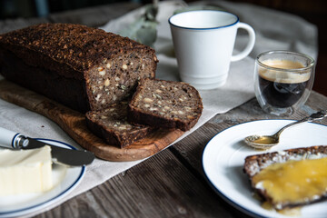 Sliced homemade traditional Danish sourdough rye bread on wooden board, cup of milk, coffee, butter and bread slice  with butter and honey on white and blue plates on wooden background.