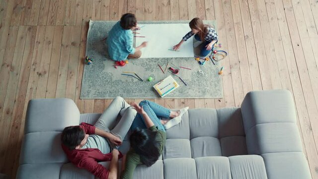 Creative activity. Top view of two little kids, brother and sister drawing with pencils, lying on the floor while young parents relaxing on the sofa