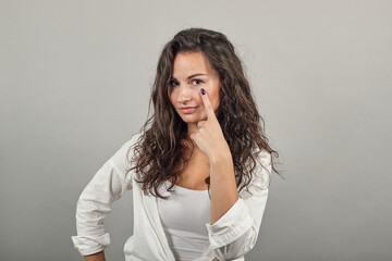 Contact lens on index finger, pointing to eye with hand, thinking, having sensitive eyes, touching herself face by forefinger. Young attractive woman, dressed white blouse, brown eyes, curly hair
