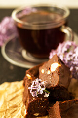 A piece of homemade chocolate cake on crumpled craft paper, with nuts on a black texture table. A cup of tea and lilac flowers decorate a delicious tea party.