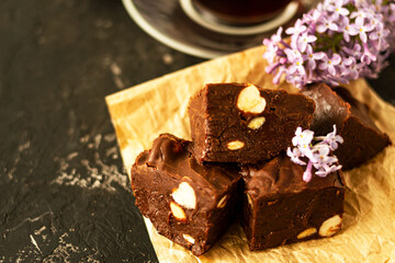 A piece of homemade chocolate cake on crumpled craft paper, with nuts on a black texture table. A cup of tea and lilac flowers decorate a delicious tea party.